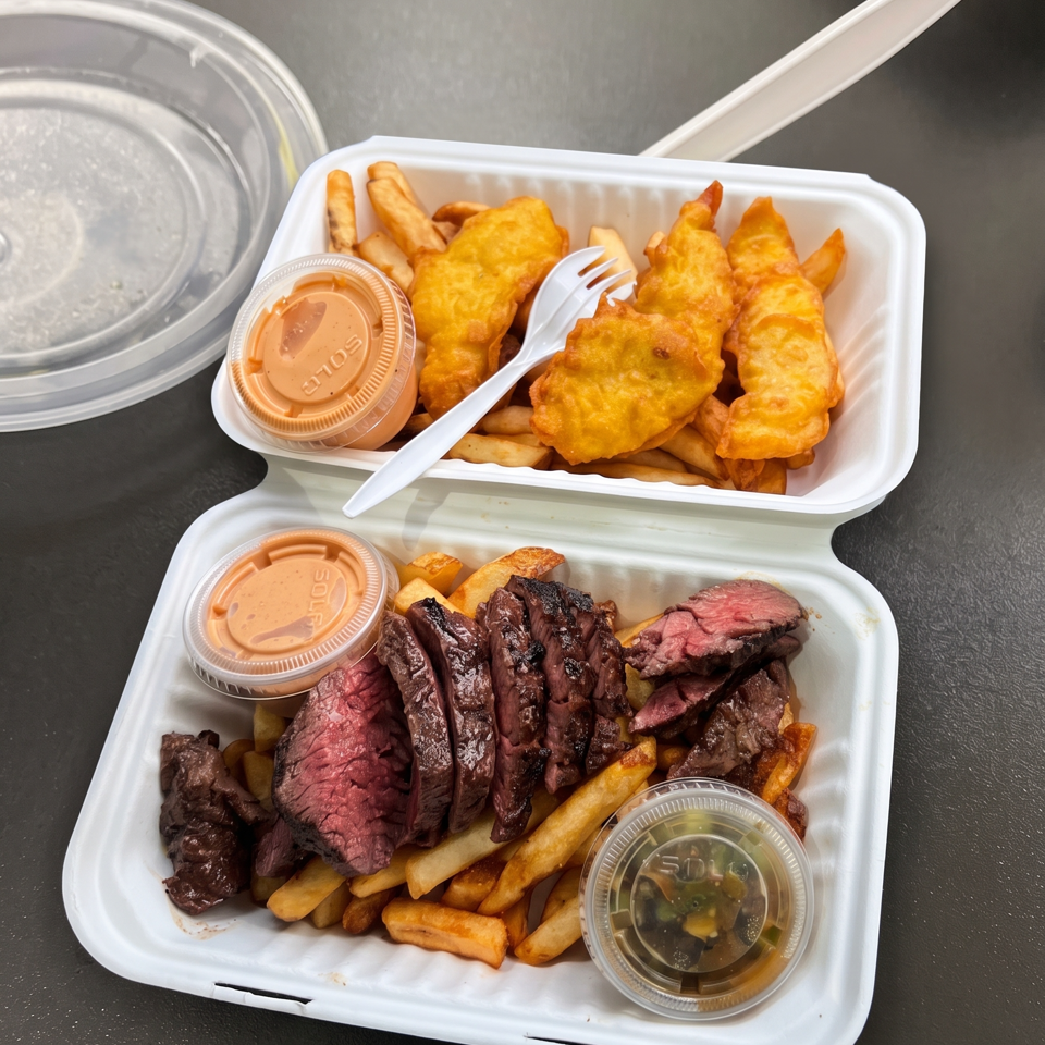 Takeout containers hold fried chicken with fries and sliced grilled steak, accompanied by dipping sauces and a plastic fork.