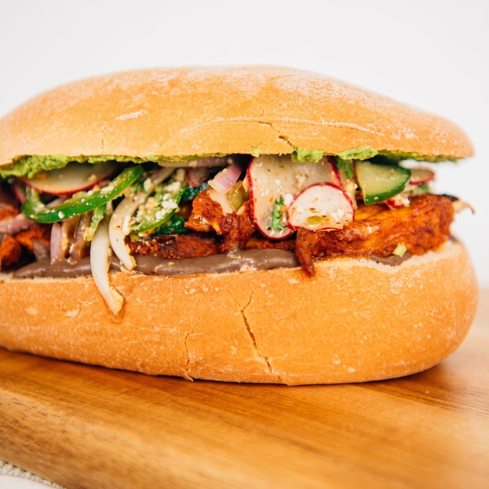 Close-up of a loaded sandwich on a toasted bun with grilled vegetables, onions, cucumber, radishes, and creamy sauce.