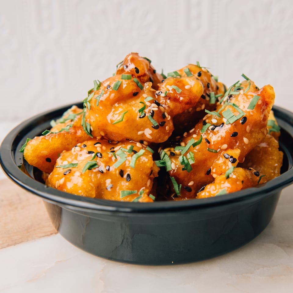 Crispy orange-glazed bites topped with black and white sesame seeds and chopped green onions in a black bowl.