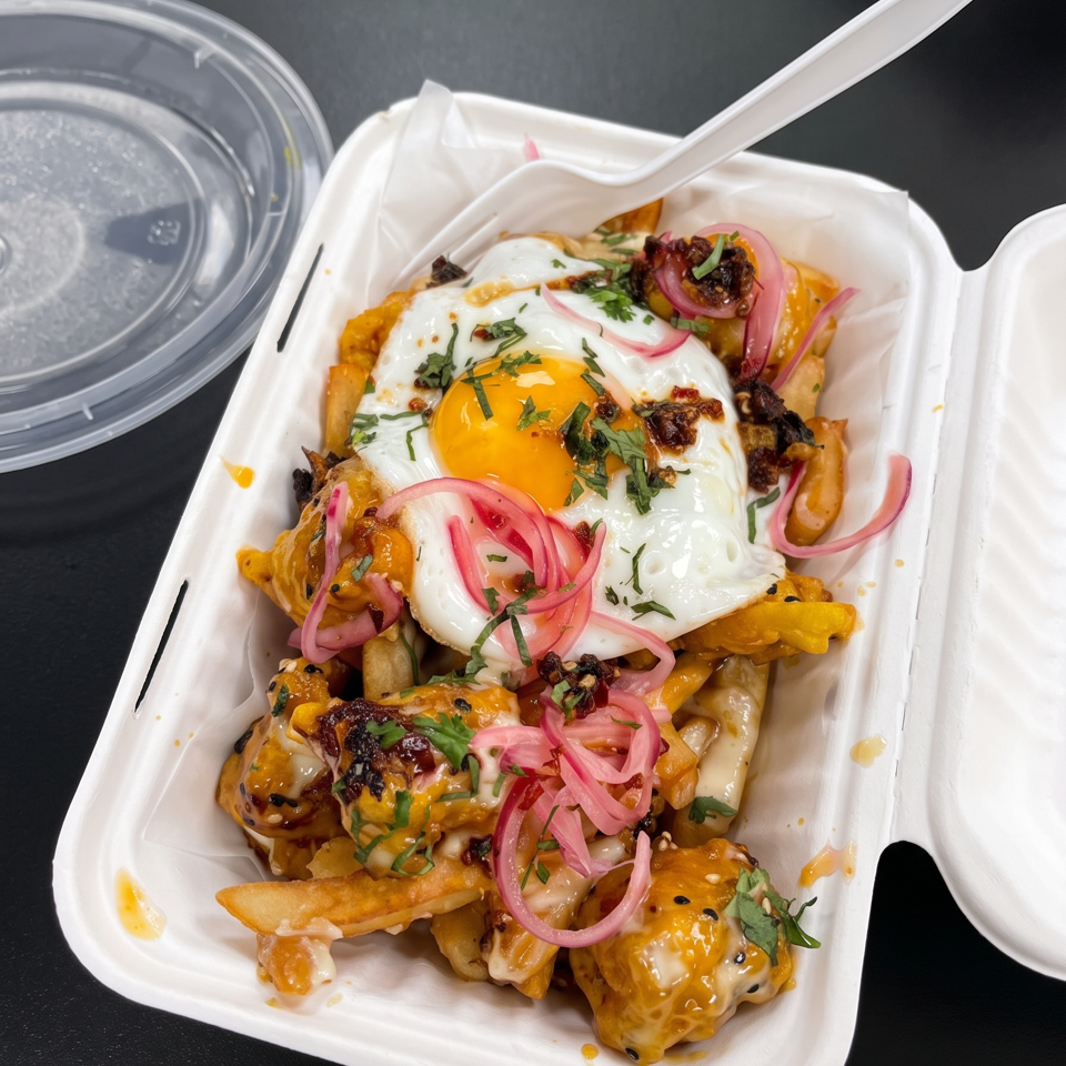 Loaded fries topped with a fried egg, cheese sauce, pickled onions, herbs, and crispy garnishes in a takeout container.